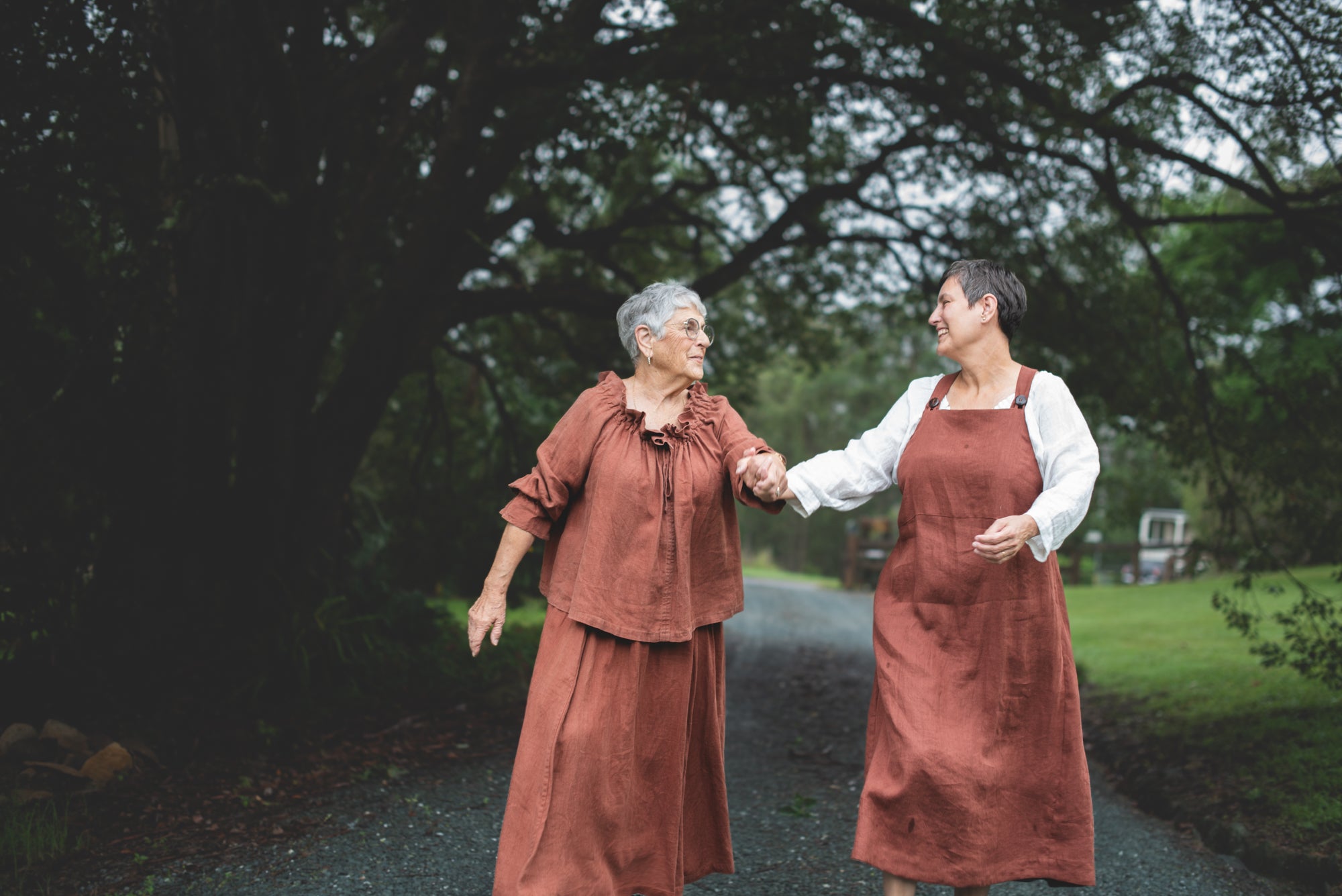 Two women in matching brown linen outfits walking together on a path with trees in the background
